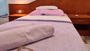 Una cama bien hecha con una manta morada, un cabecero de madera, una mesita de noche con una lámpara y un pequeño mueble auxiliar con un control remoto.