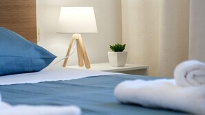 Un lit bien fait, avec une literie bleue, un oreiller blanc et une serviette blanche enroulée. On y trouve une petite table de chevet avec une lampe, une plante en pot et un présentoir en bois.