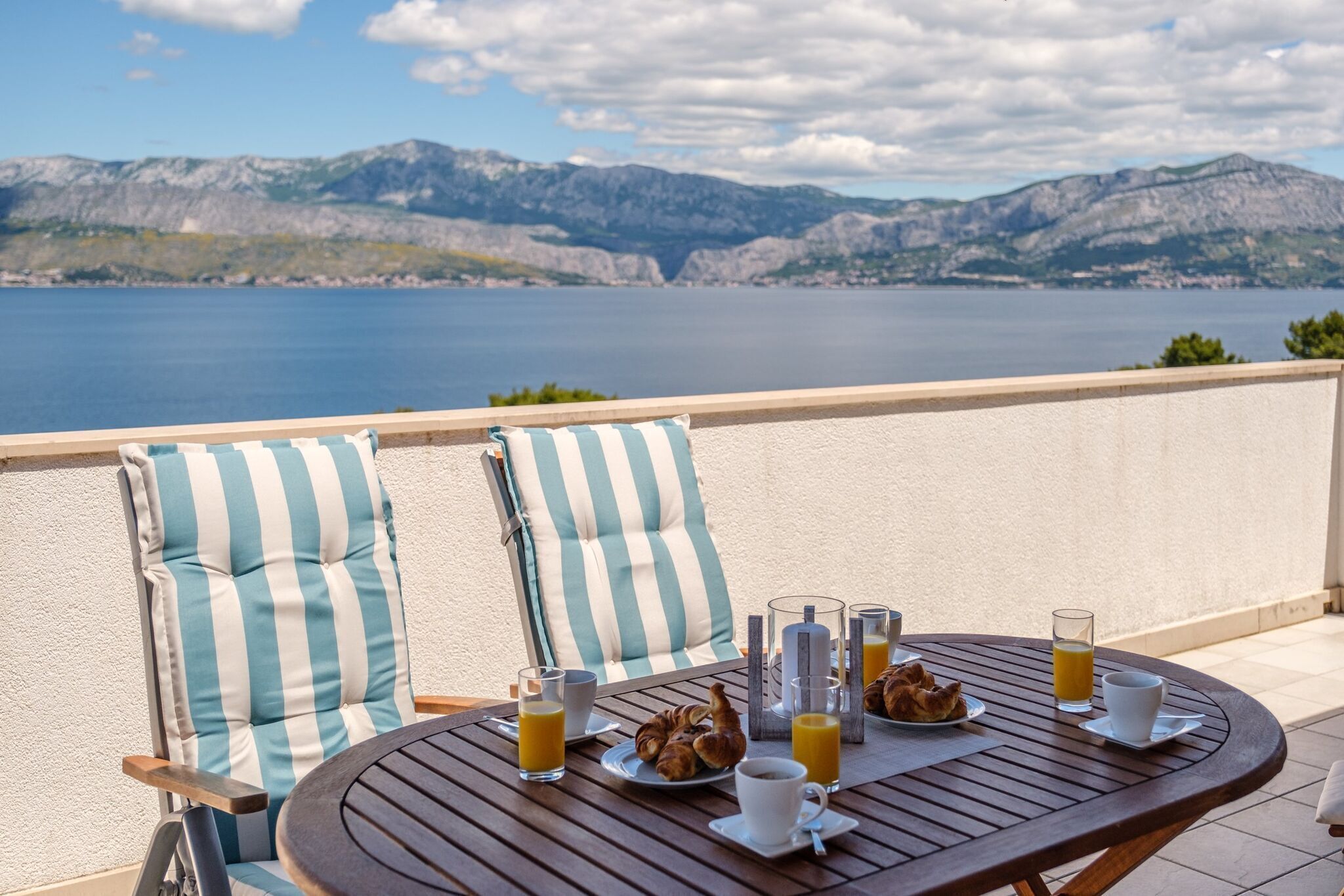 Ein Esstisch mit Frühstücksgebäck, ein Stuhl mit gestreiften Kissen und eine herrliche Aussicht auf Berge und Wasser.