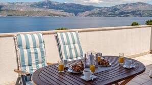 Une table à manger avec des aliments pour le petit-déjeuner, une chaise avec des coussins à rayures et une vue panoramique sur les montagnes et l’eau.