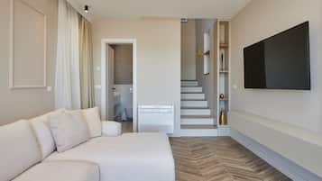 Une chambre d’hôtel moderne dotée d’un canapé, d’un téléviseur à écran plat fixé au mur, d’un parquet à motifs en chevrons et d’un escalier menant à un niveau supérieur.