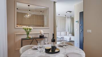 Un espace repas moderne avec une table ronde dressée pour quatre personnes, une bouteille de vin et un vase rempli de fleurs. On aperçoit un grand miroir et un canapé à l’arrière-plan.