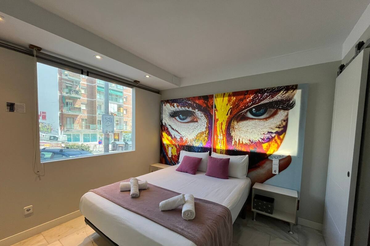 Una habitación de hotel moderna con una cama grande, una ventana con vistas a la ciudad y un cuadro abstracto llamativo en la pared.