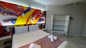 A modern hotel room with a large bed, a headboard with a colorful abstract design, a white bedside table, a built-in shelf unit, and a marble floor.