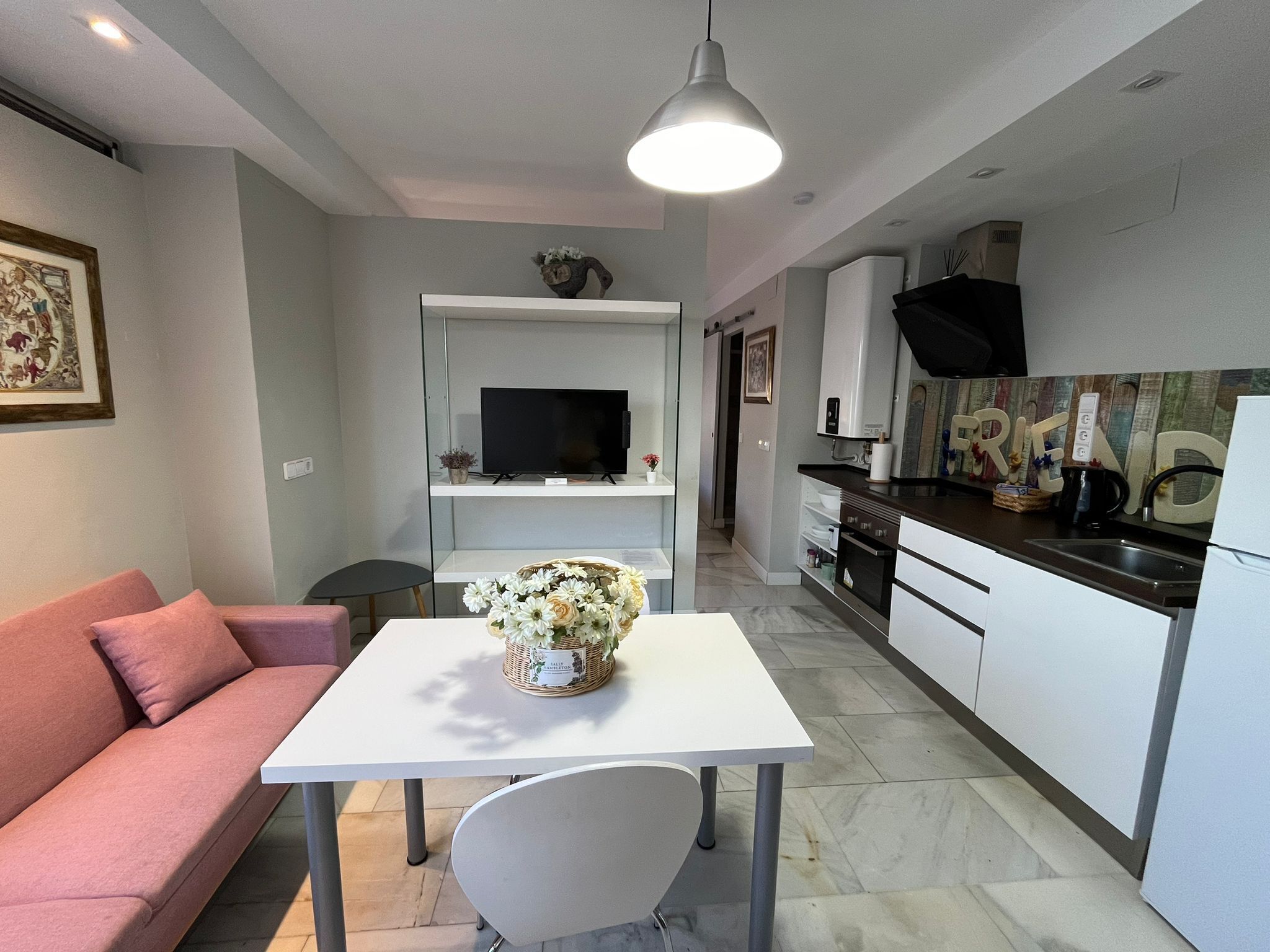 Una cocina moderna con armarios blancos, encimera negra y horno empotrado. En la sala de estar, hay una mesa de comedor con una silla blanca y un sofá rosa. Un televisor de pantalla plana está montado sobre la mesa.