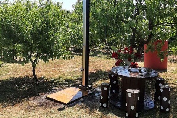 Un espace extérieur aménagé avec une table et des tabourets en bois, entouré d’arbres et agrémenté d’un jardinier rouge rempli de fleurs.
