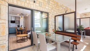 Un salon moderne avec une table à manger en verre, des chaises en osier et un banc. On peut apercevoir la cuisine à travers une porte en verre, et une grande fenêtre laisse entrer la lumière naturelle.