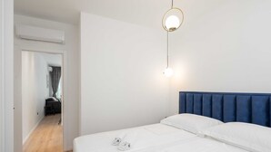 Une chambre d’hôtel avec un lit, une tête de lit bleue, un luminaire suspendu et une vue sur un couloir avec une porte donnant sur une autre chambre.
