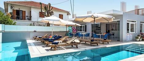 A swimming pool with lounge chairs and umbrellas, a dining area with tables and chairs, and a building with a tiled roof.