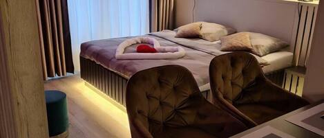 Une chambre moderne avec un lit, deux fauteuils rembourrés, une table de chevet et une fenêtre avec des rideaux.