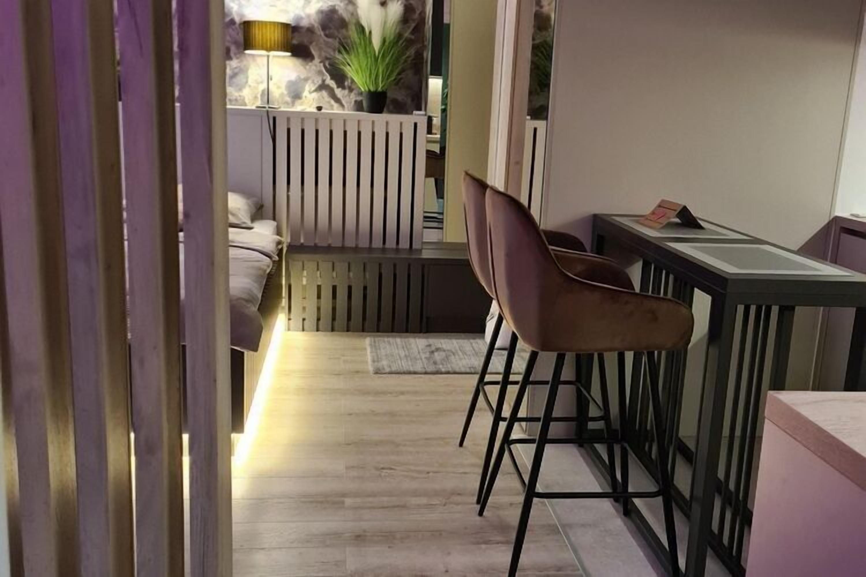 Une chambre moderne avec un plancher en bois, un coin bar avec des tabourets, un lit avec une tête de lit blanche et une plante en pot.
