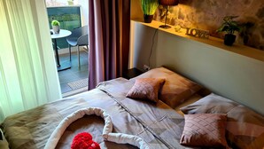 Une chambre à coucher avec un lit, des oreillers, un coussin en forme de cœur rouge, une petite table avec une plante et une étagère avec une décoration représentant le mot « AMOUR » et d’autres plantes.
