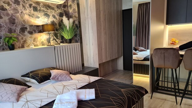 Une chambre moderne avec un mur en pierre, un lit avec une literie à motifs géométriques, une table de chevet avec une lampe, et un petit coin salon avec un fauteuil et un saladier de fruits.