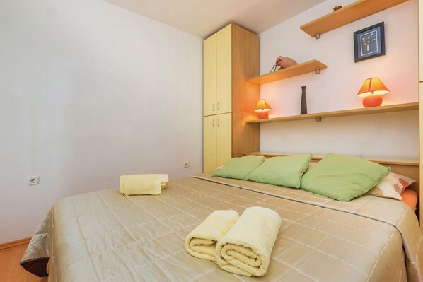 Una cama bien hecha con almohadas verdes y dos toallas enrolladas, un armario de madera y dos lámparas fijadas a la pared.