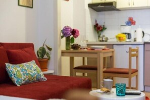 A cozy room with a red sofa, a small table with a cup and a remote control, and a kitchen area with flowers and a coffee maker.