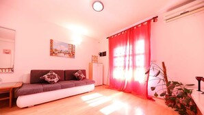 A living room with a sofa, a small table, a potted plant, and a large window with red curtains.