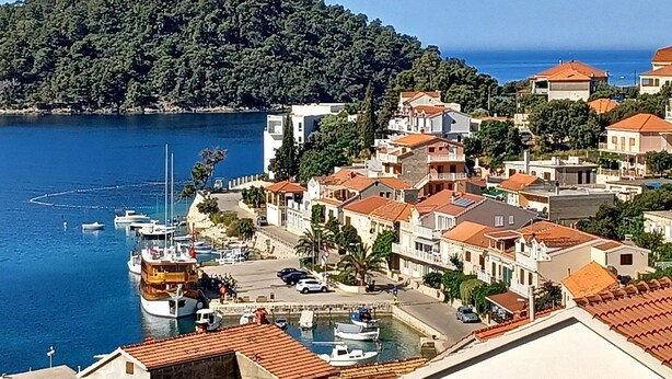 Un pueblo costero con edificios a lo largo del malecón, un puerto deportivo con barcos y una colina al fondo.