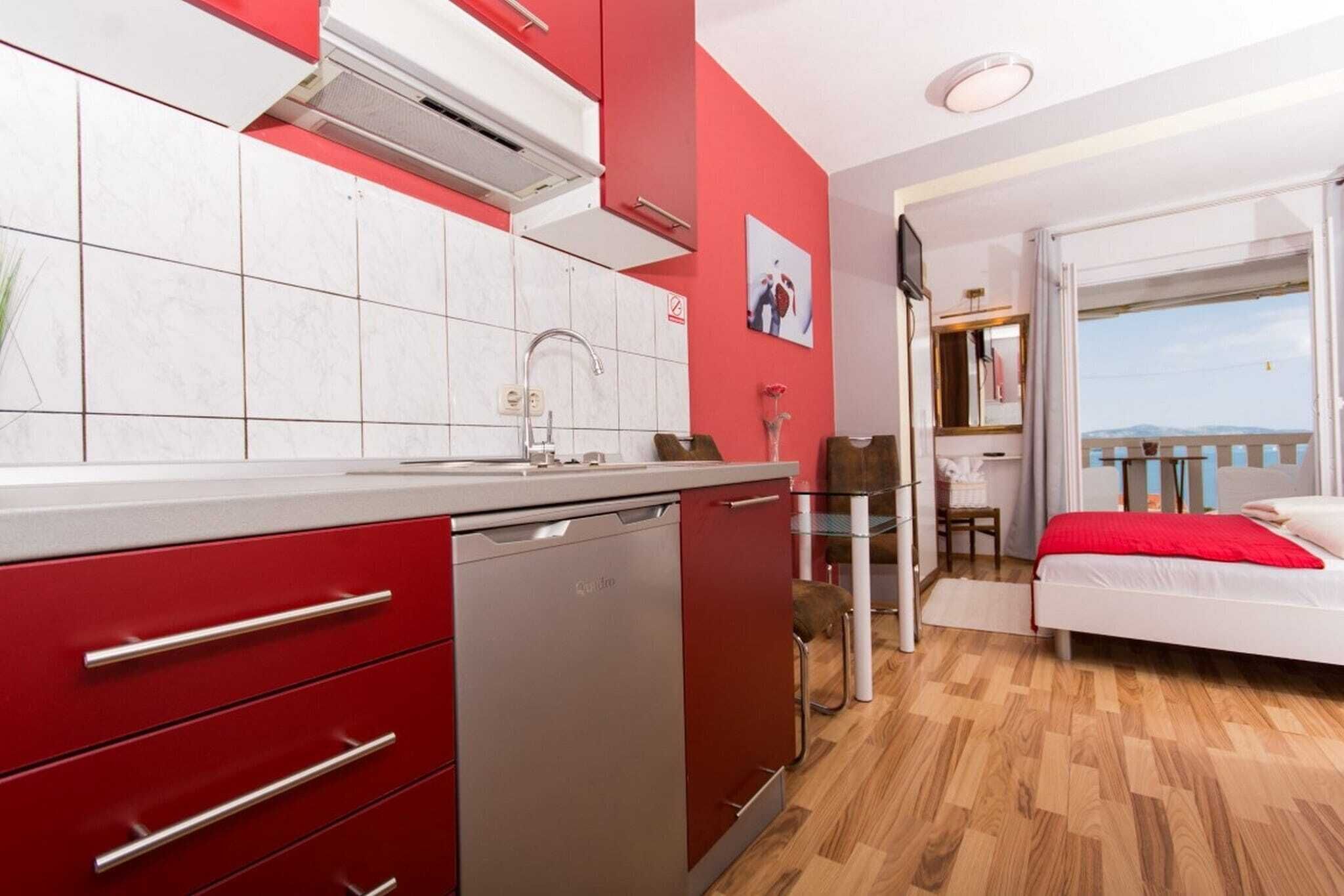 Een compacte keuken met rode keukenkastjes, een vaatwasser en een betegelde spatwand. Grenzend aan de keuken bevindt zich een klein eethoekje met een tafel en stoelen. Een bed met een rode deken is in de kamer zichtbaar.