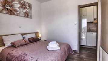 A bedroom with a bed, pillows, a painting, and a view into the kitchen.