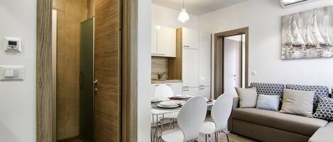 A modern living room with a sofa, dining table, and chairs. There is a wooden door and a white wall with a painting.