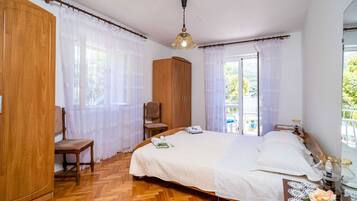 A bedroom with a wooden wardrobe, a chair, a bed with white linens, and a window with a view of greenery.