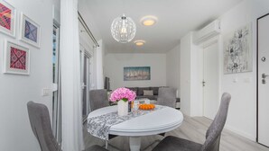 A dining area with a round table, grey upholstered chairs, a white tablecloth, a vase with pink flowers, and a bowl of oranges.