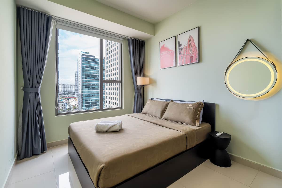 A hotel room with a large window, a bed with a beige bedspread, a round mirror, and two framed pictures on the wall.