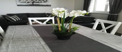 Un comedor con una mesa preparada para dos personas, un camino de mesa negro, sillas blancas y una planta en maceta con flores blancas.