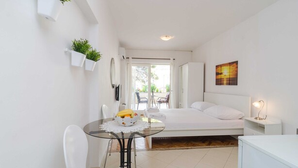A compact bedroom with a bed, bedside table, and a small table with a fruit bowl. There are white chairs, a glass table, and a view of the outdoors through a door. - House Apartments Ariva - Studio Apartment with Balcony (2 Adults) - A2 (Dubrovnik)