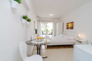 A compact bedroom with a bed, bedside table, and a small table with a fruit bowl. There are white chairs, a glass table, and a view of the outdoors through a door.