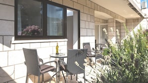 An outdoor patio with wicker furniture, a table with a bottle and glass, and a window overlooking a garden.
