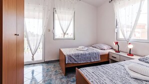 Ein Schlafzimmer mit zwei Betten, einem Holzschrank, einem Fenster mit durchscheinenden Vorhängen und einem Nachttisch mit Lampe.