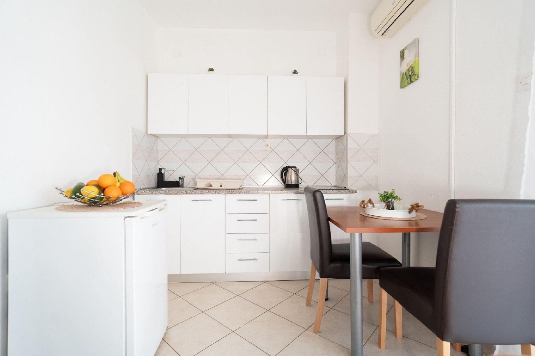 Une cuisine compacte avec des meubles blancs, une petite table à manger avec deux chaises et un panier de fruits posé sur le comptoir.
