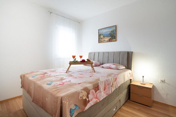 Une chambre à coucher avec un lit, une table de chevet, une commode, un tableau accroché au mur et une fenêtre avec des rideaux.