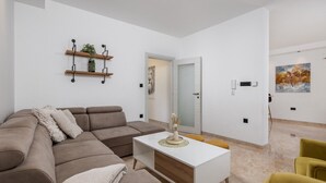 A modern living room with a sectional sofa, a white coffee table, and a yellow armchair. There are two wooden shelves with plants and a painting on the wall.