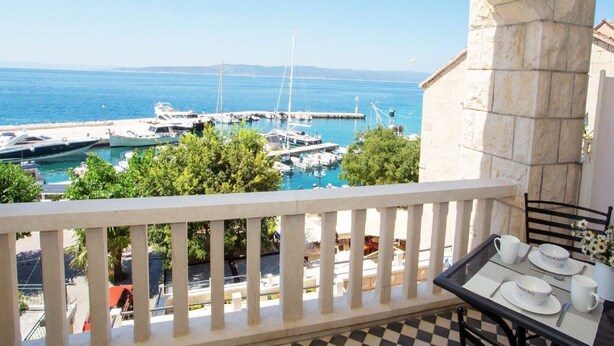 A balcony with a view of a marina and boats, a table with white plates and cups, and a stone building. - Apartments & Room Vesna - Superior Studio Apartment with Sea View (Brela)