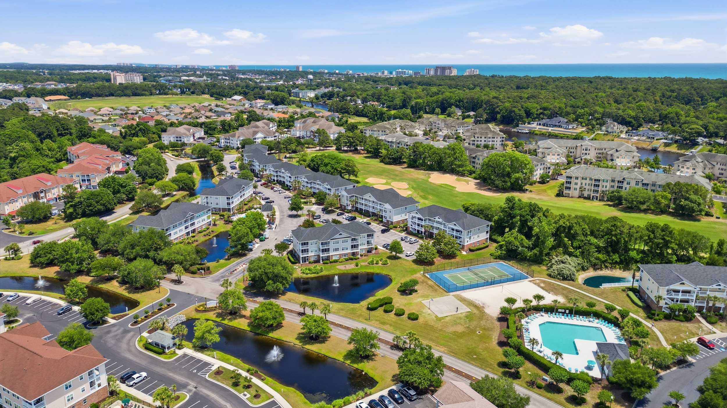 A residential area with apartment buildings, a tennis court, a swimming pool, and a lake.