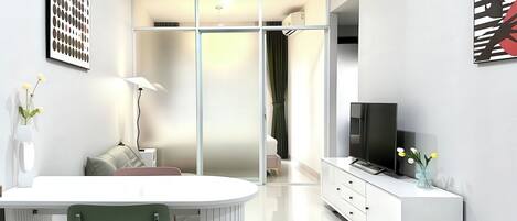 A modern, well-lit living room with a white sofa, a dining area with a table and chairs, a TV on a white cabinet, and artwork on the walls.