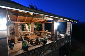 An outdoor patio area with wooden furniture and potted plants, featuring a stone wall and a wooden roof structure.