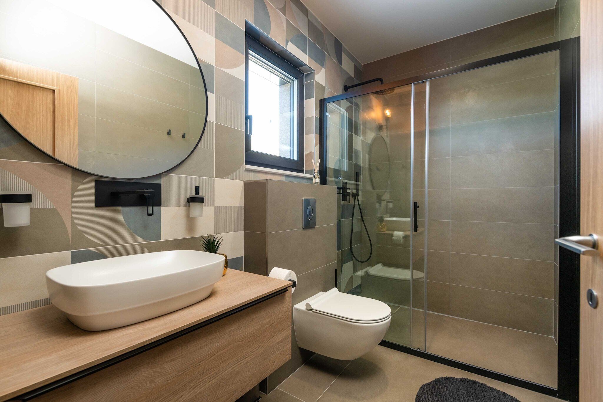 A modern bathroom with a large oval mirror, a white sink, a toilet, and a glass-enclosed shower area.