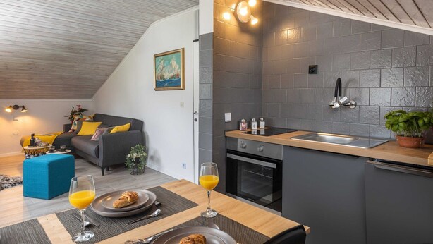 Une cuisine moderne dotée d’un four encastré et d’un évier, un coin repas avec une table dressée pour deux personnes, et un espace de détente avec un canapé et des coussins.
