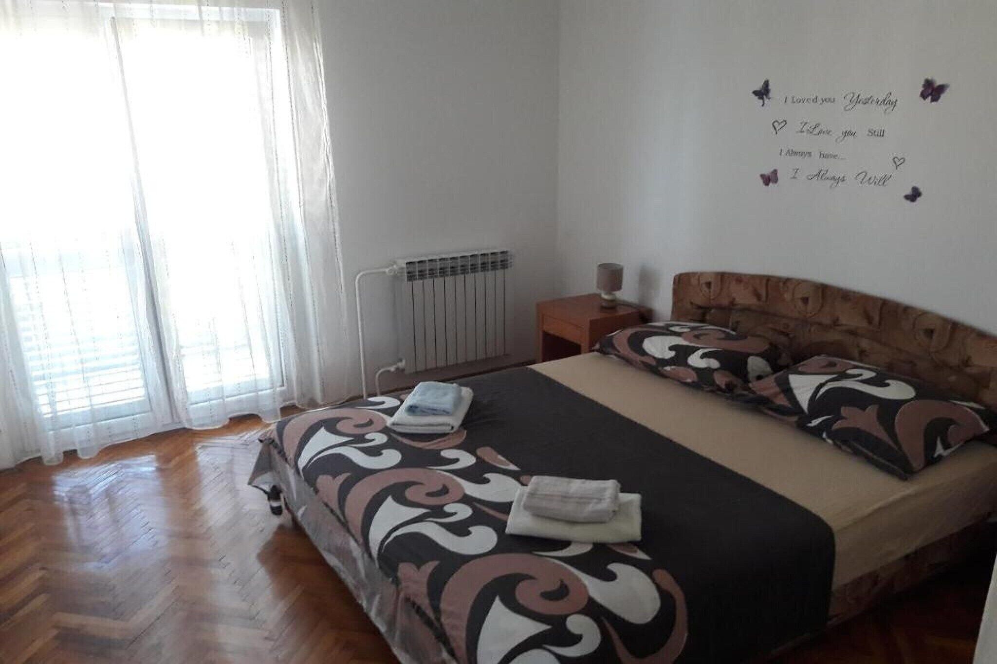 A bedroom with a bed, a nightstand, a wall clock, a radiator, and a window with curtains.