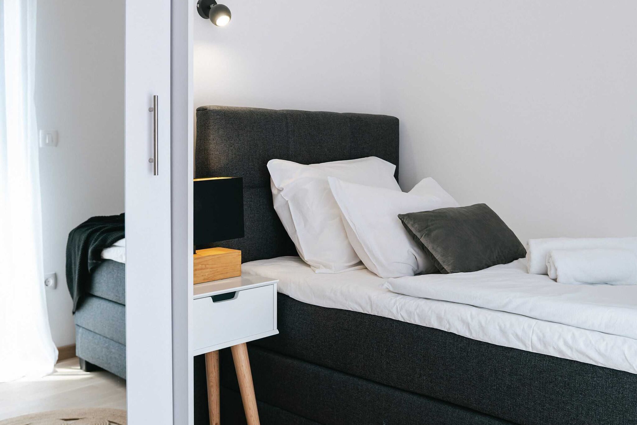 A modern bedroom with a dark grey headboard, white bedding, and a bedside table with a lamp and a box.