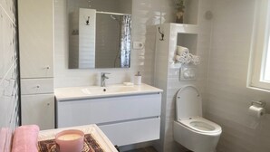 Un baño moderno con un lavamanos, espejo y retrete blancos. Hay una toalla en el suelo y una vela sobre una bandeja.