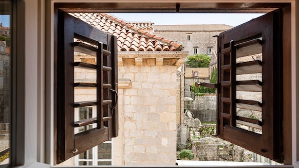 Vista a través de contraventanas de madera de un patio con edificios de piedra y techo de tejas.