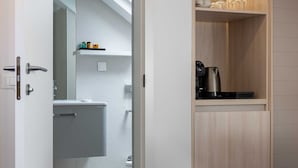 Un baño moderno con ducha, inodoro y lavamanos. Hay un estante de madera con una cafetera y un espejo pequeño.