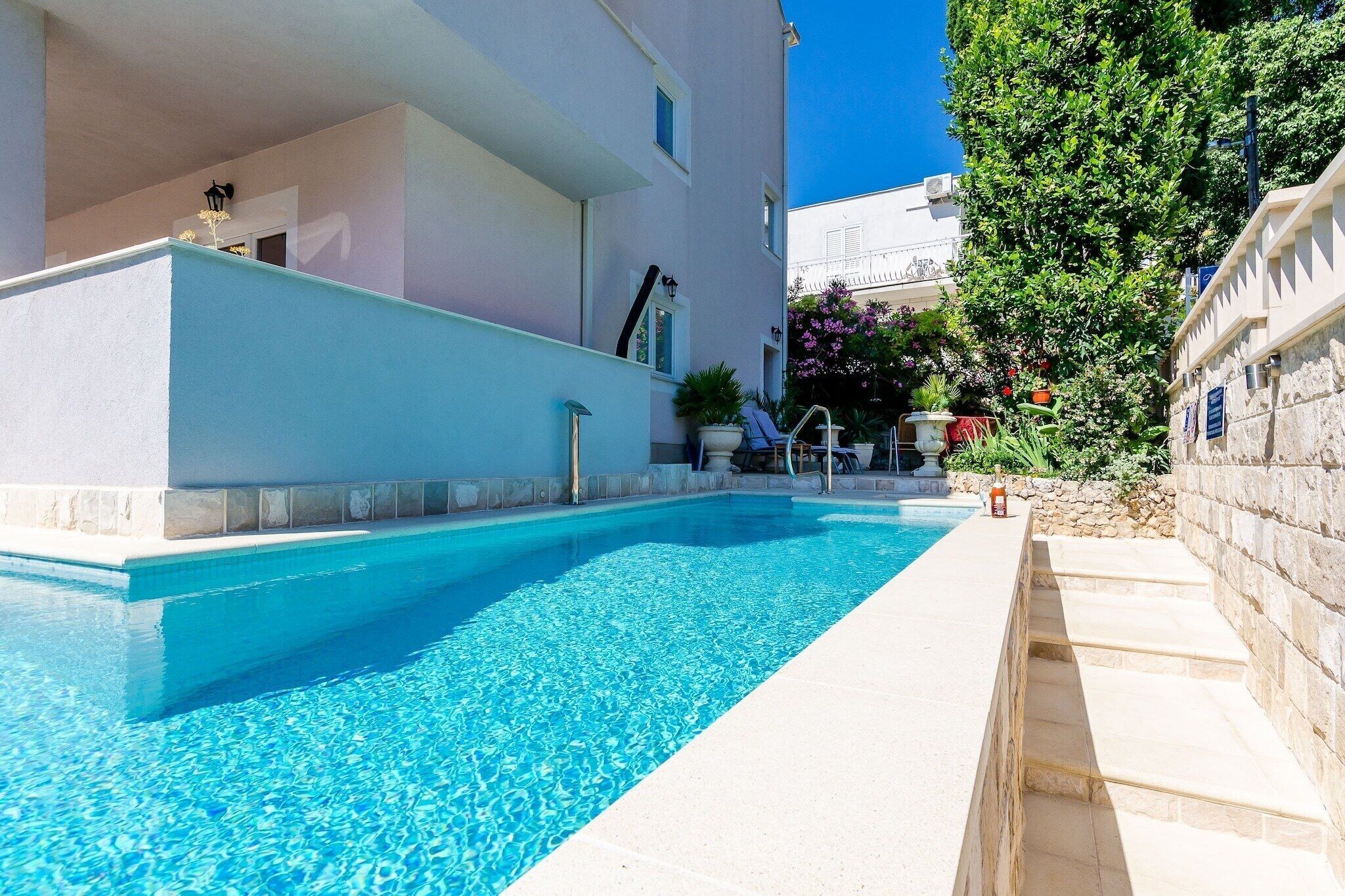 Uma área de piscina com uma piscina azul cristalina, degraus que levam à água e um edifício com fachada branca.