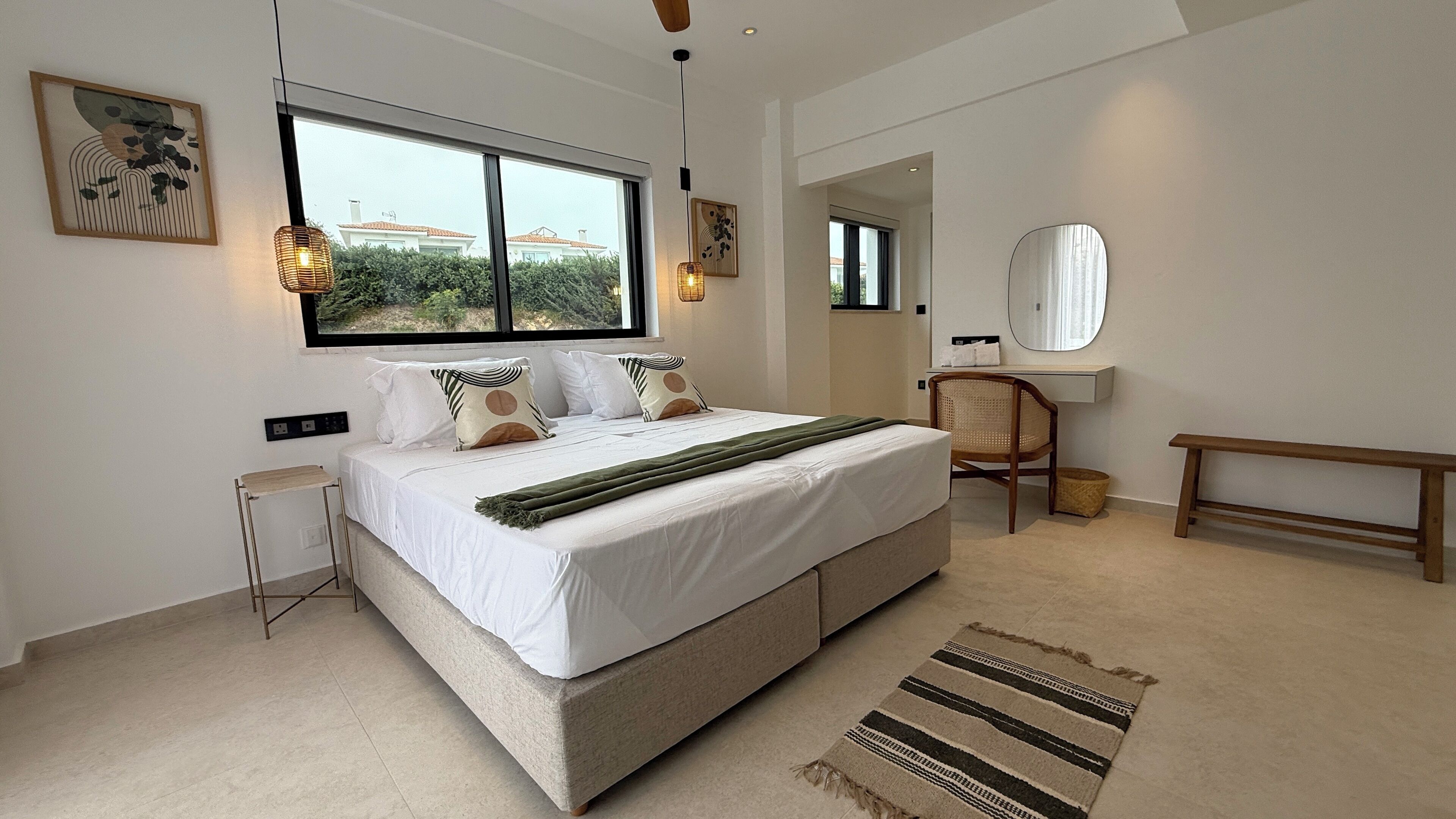 A modern bedroom with a large bed, a bedside table, a chair, a bench, a mirror, and a window with a view of houses.