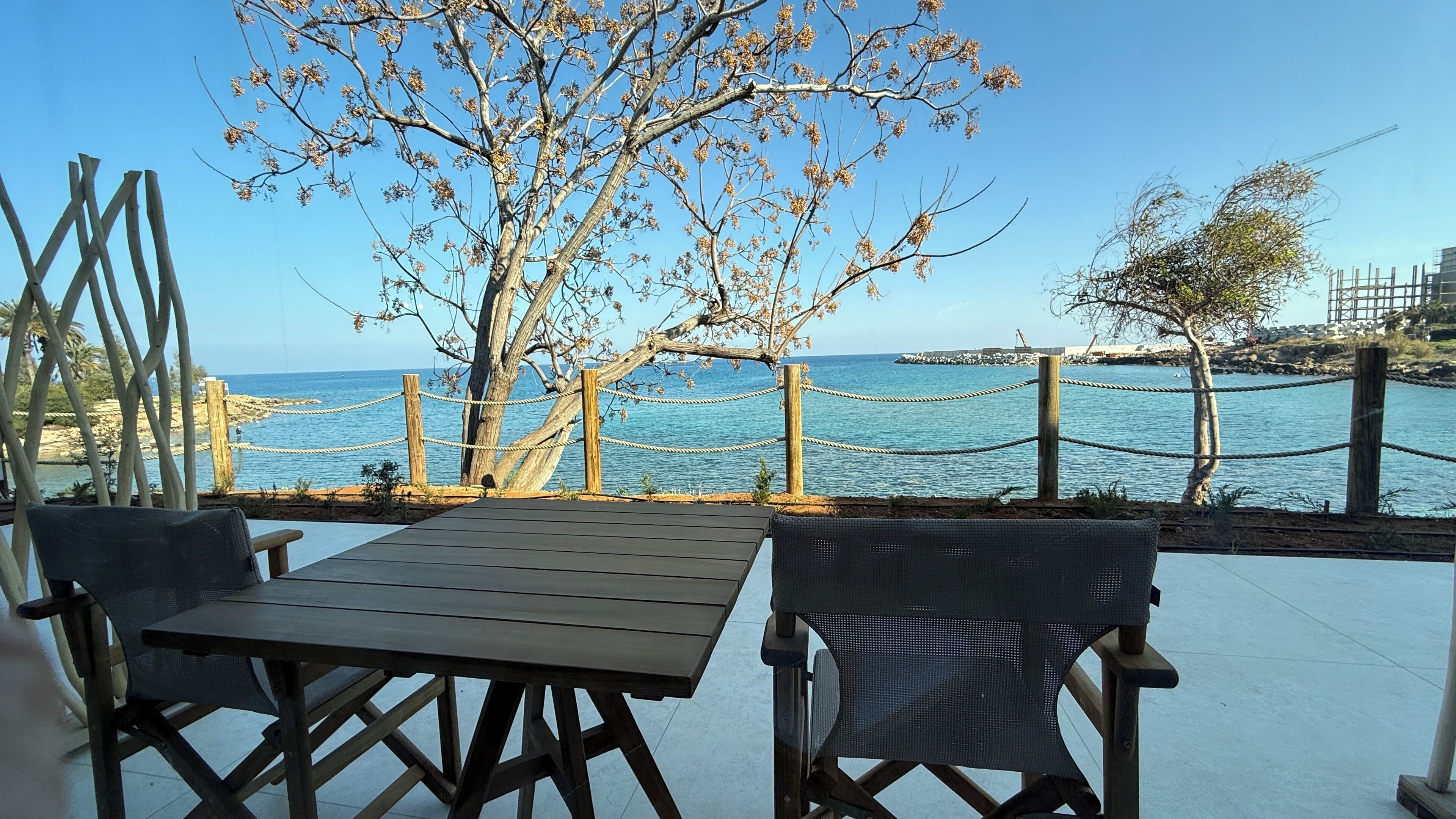 An outdoor seating area with a view of the sea, wooden tables and chairs, and a rope barrier.