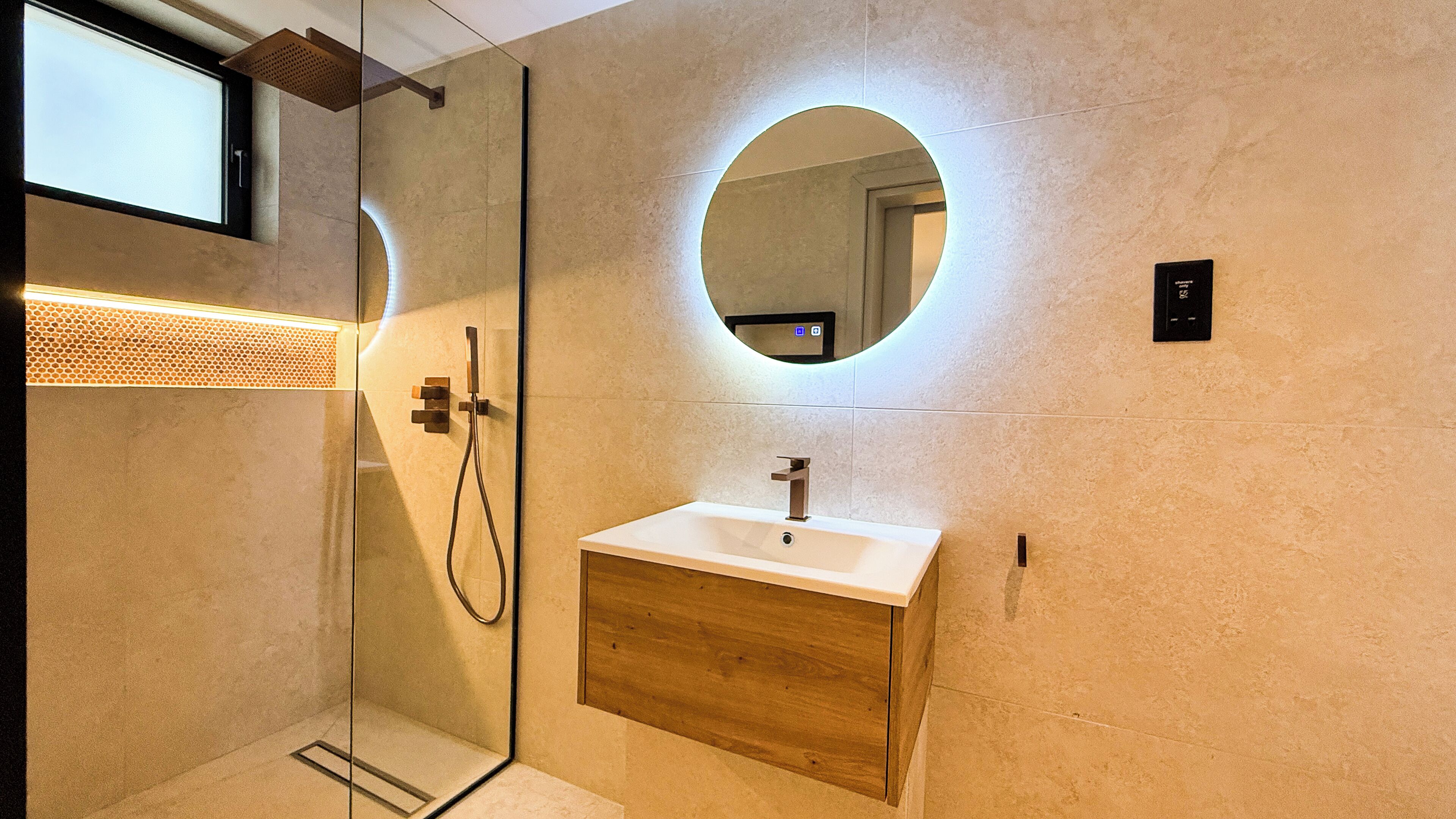 A modern bathroom with a glass-enclosed shower, a wooden vanity, and a round mirror with a light.